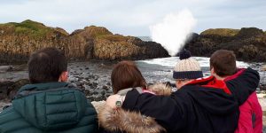 AR16 (front cover) Four people in coats standing together with arms round each other staring out at the coast.